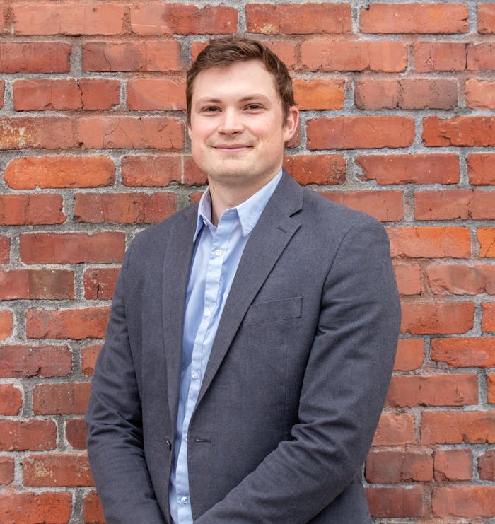 man-in-gray-suit-standing-beside-brick-wall-14634927 Smiling man in a suit posing against a brick wall, exuding confidence and professionalism.